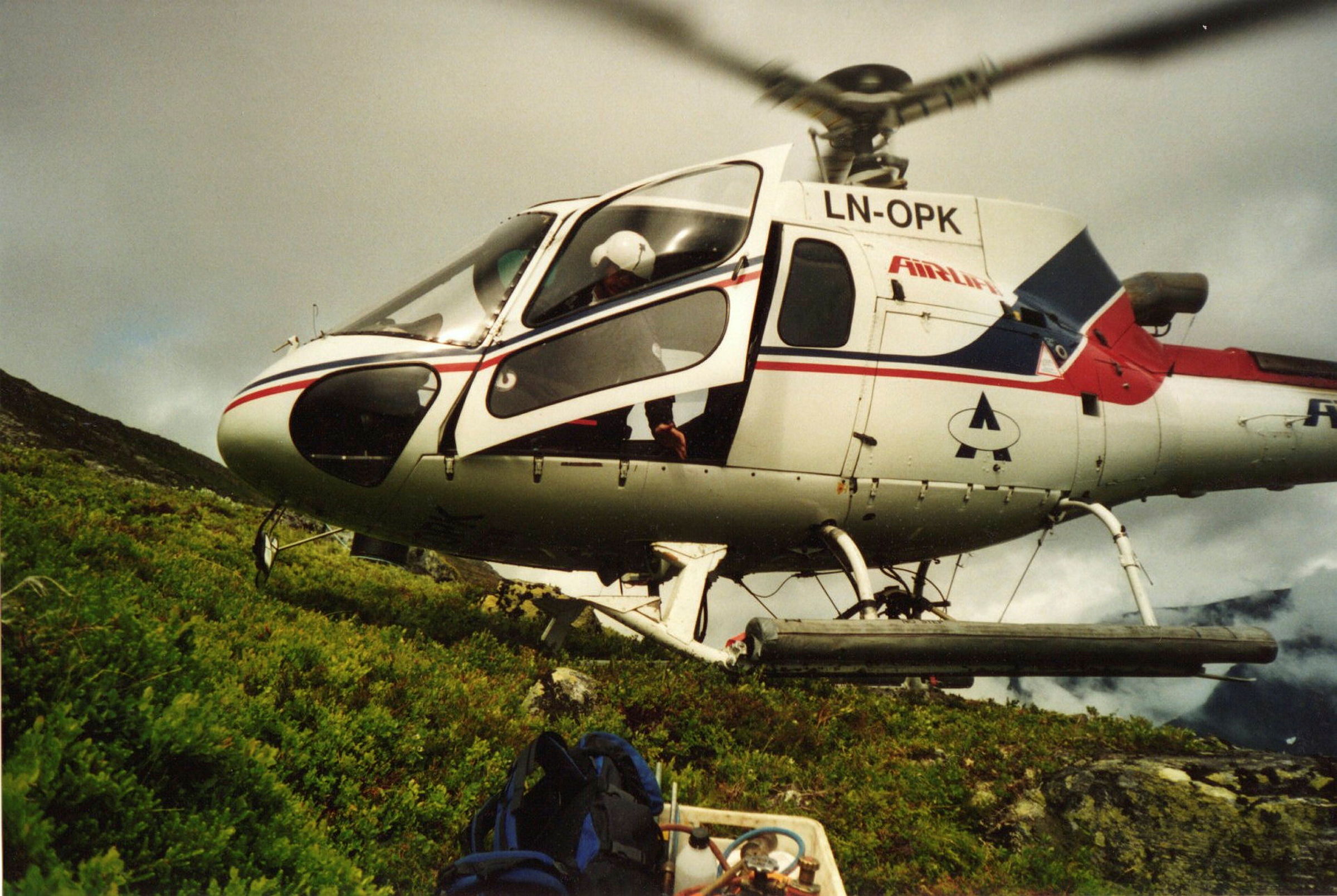 Et helikopter med snurrende propell står inntil en bakke i gress. En mann med hjelm er på vei ut døra av helikopteret.