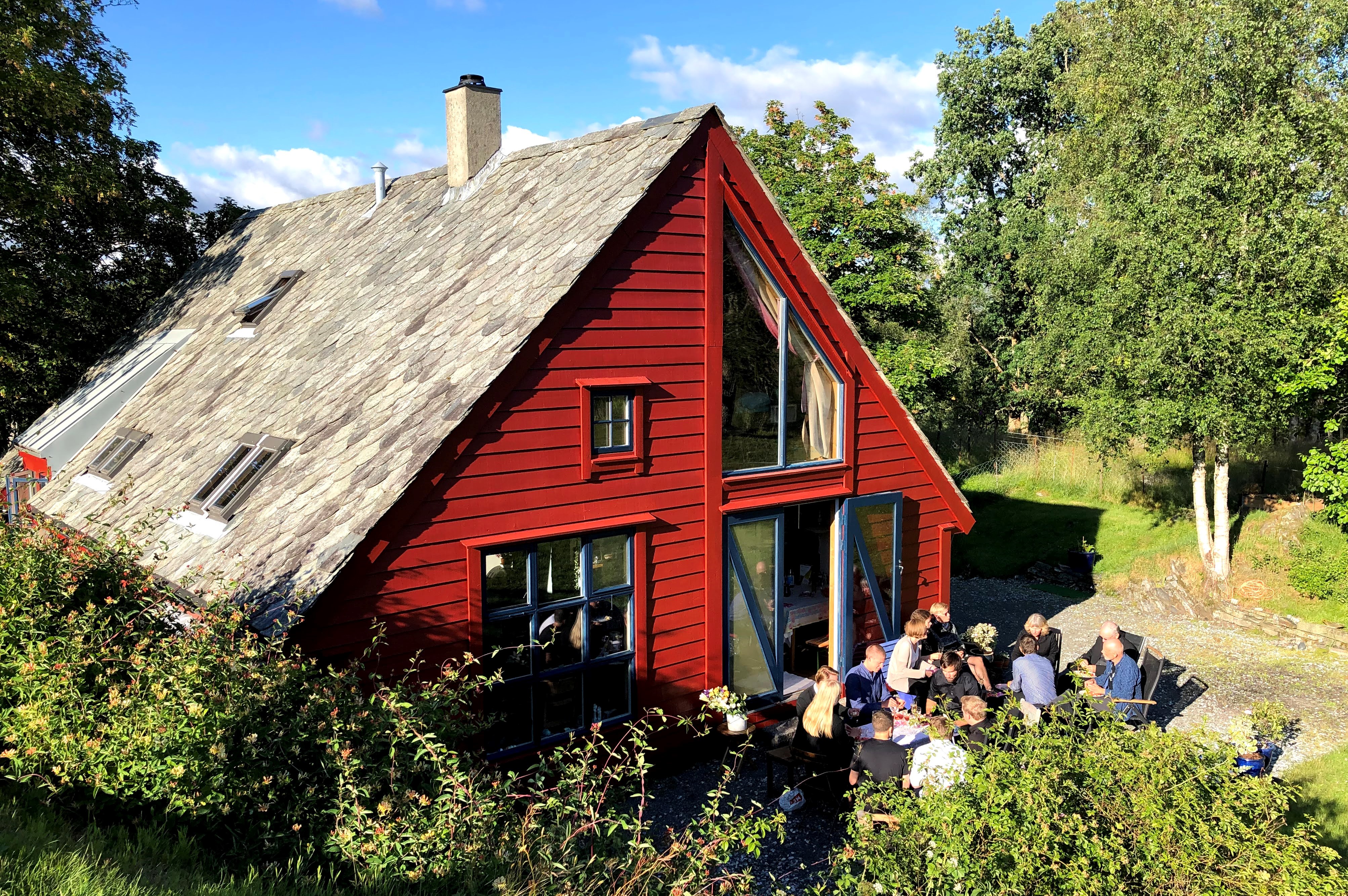 Bilde av et rødmalt enebolig med mennesker utenfor i hagen, omgitt av grønne vegetasjon.