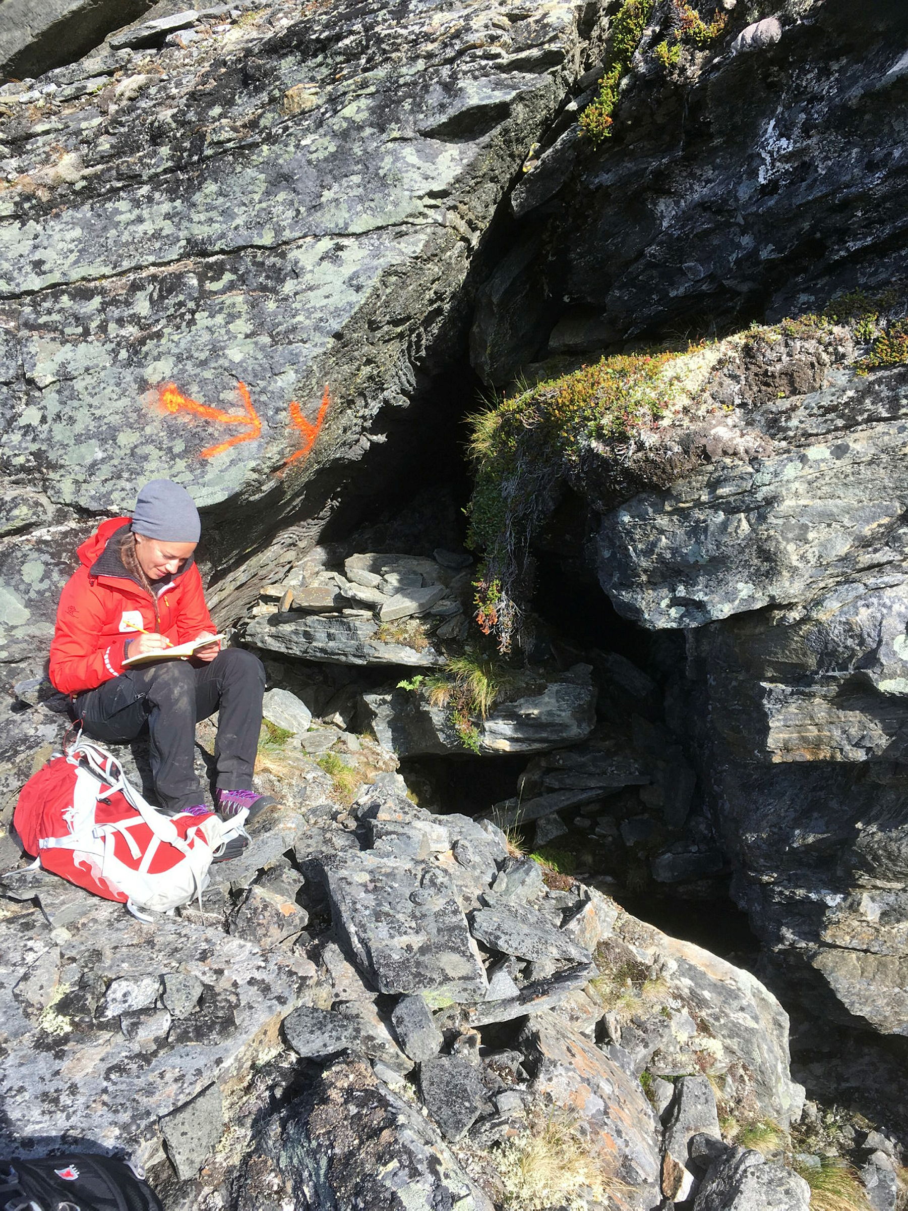 Person i rød NVE jakke sitter foran en fjellsprekk og noterer i notatblokk.