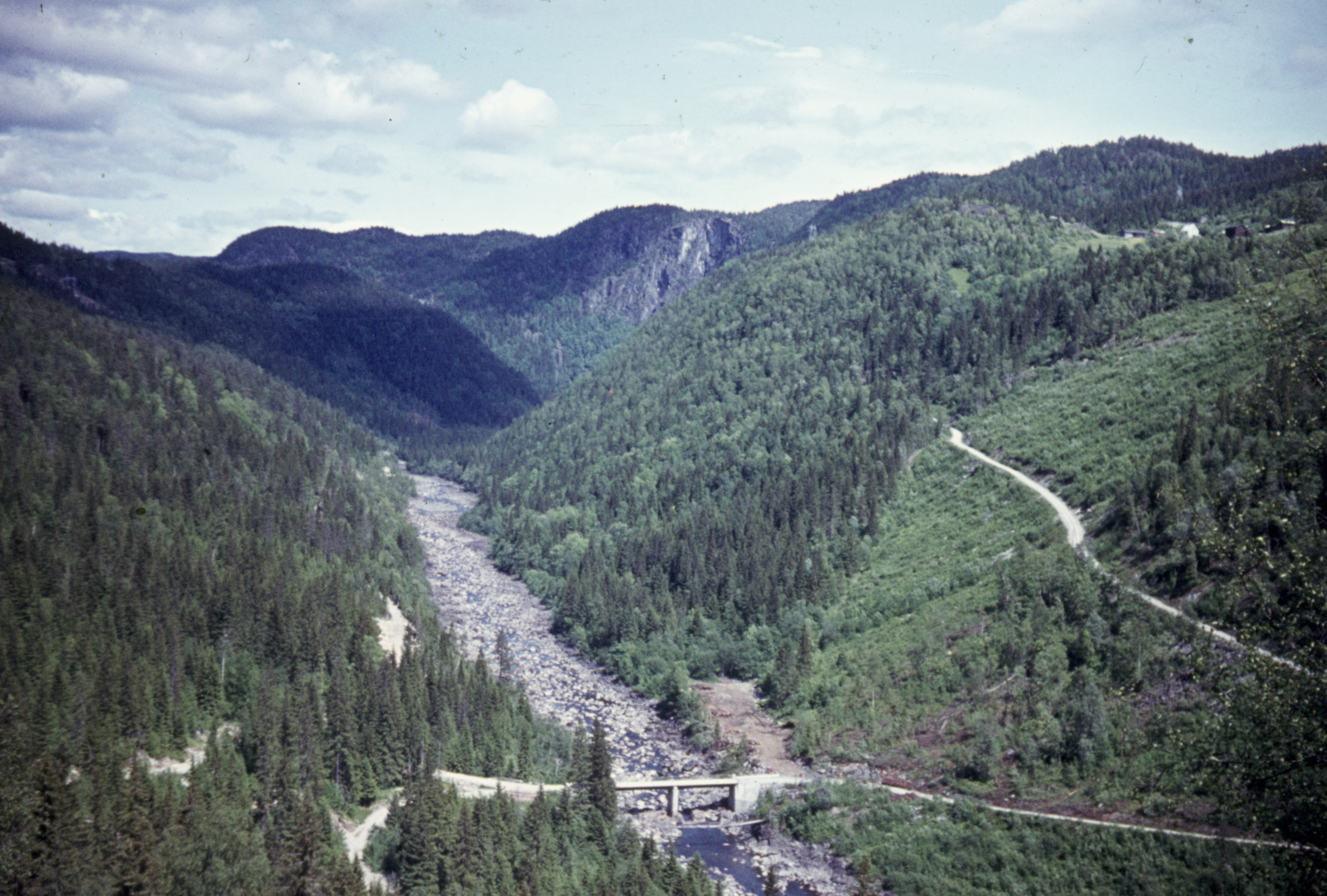 Tørrlagt elv med lite vannføring som renner gjennom en dal. Bilvei og bru som krysser elva. 