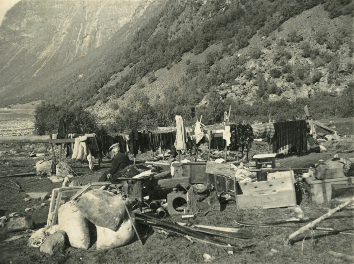 Blant ruinrester foran en fjord med høye fjell henger det igjen klesstativer i tre med klær til tørk.