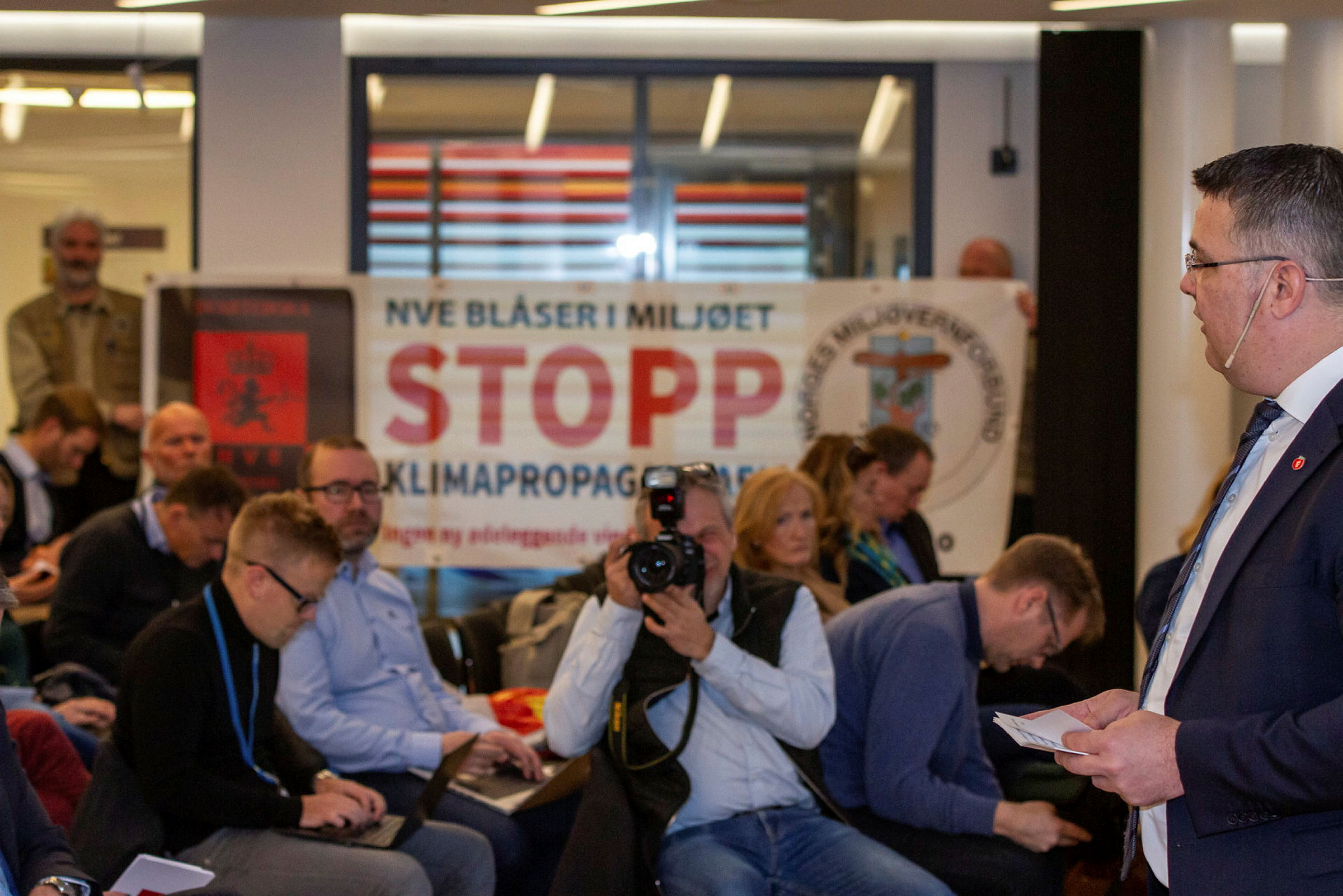 En grupper personer ser på mann som snakker. I bakgrunnen banner med budskapet: NVE blåser i miljøet. Stopp klimapropagandaen.