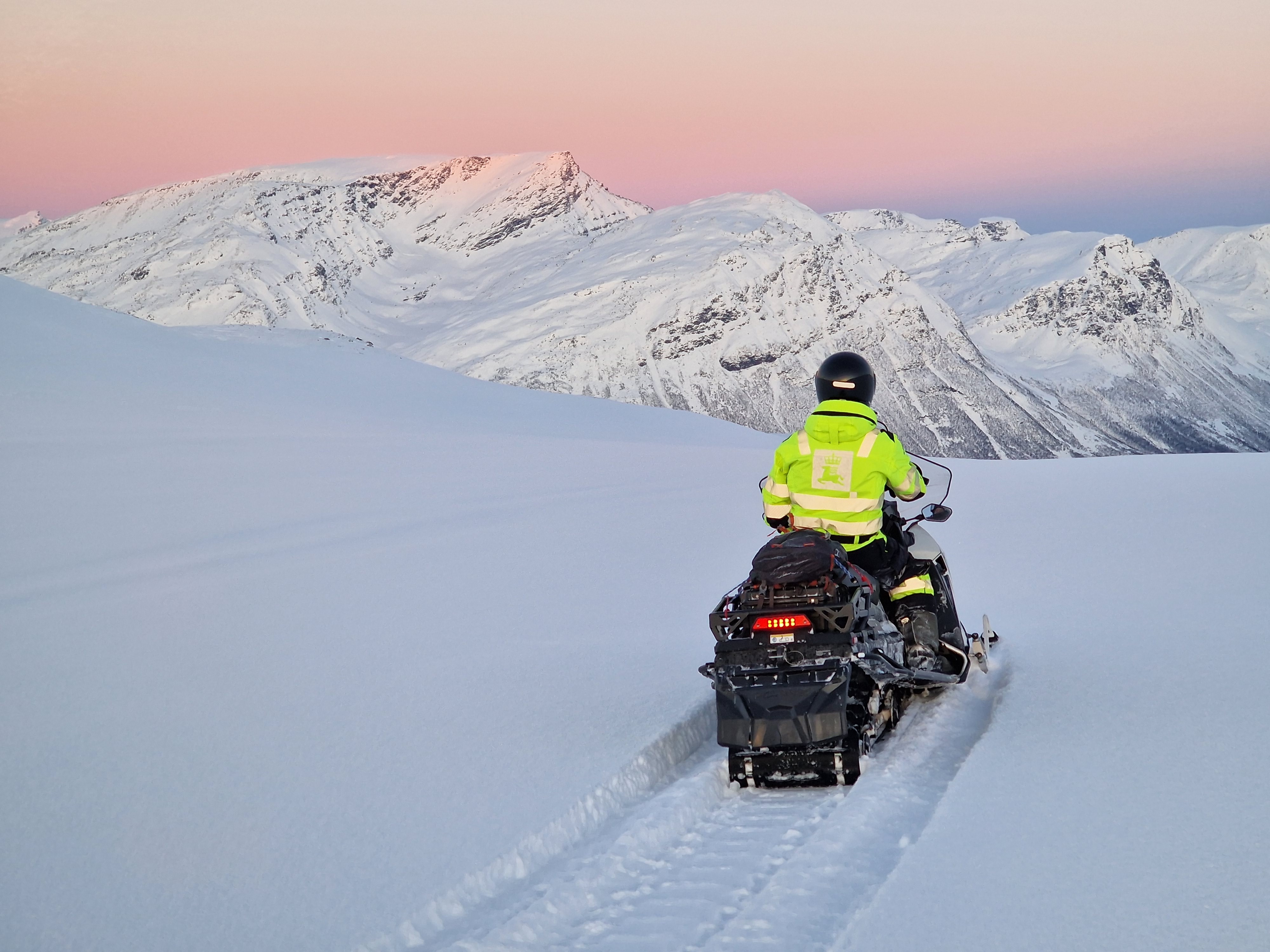 En person i neon arbeidsklær sitter med ryggen til og kjører en snøscooter i fjellet. I bakgrunnen er det snødekte fjell og en rosa himmel.
