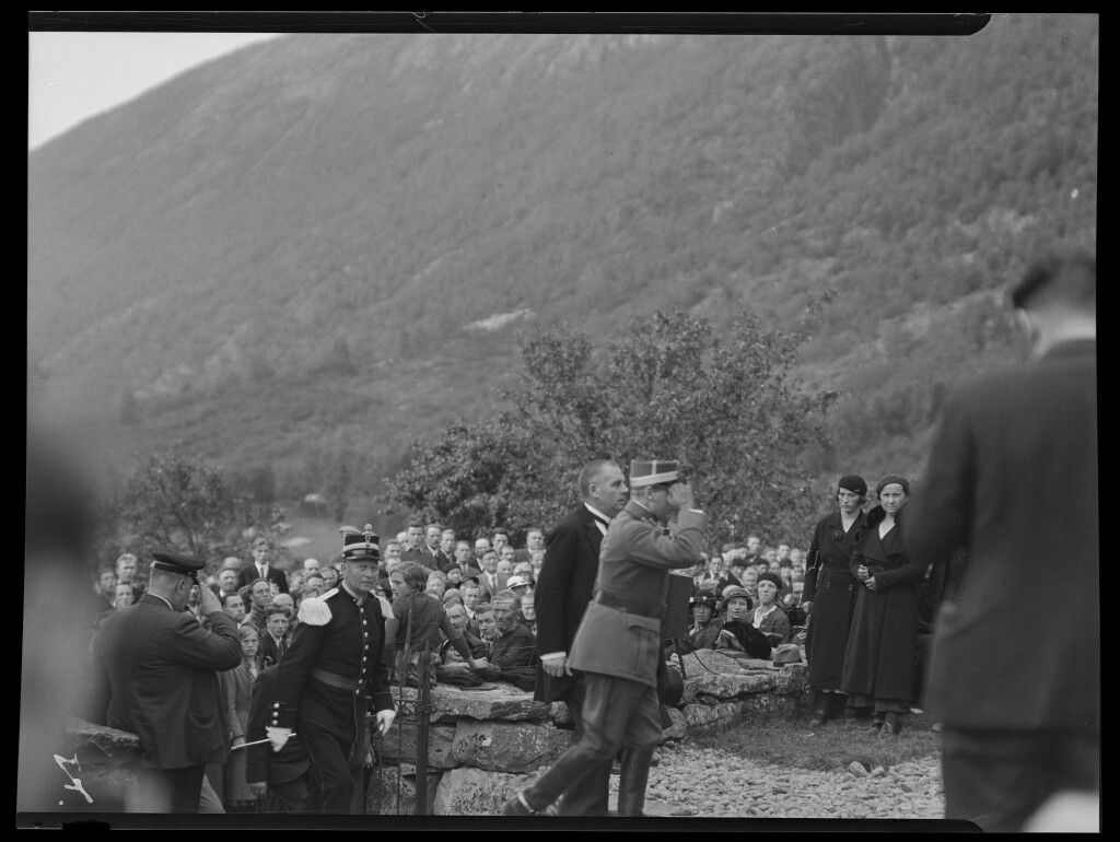 Et svart-hvitt bilde av uniformerte menn som går på rekke. Rundt dem står en stor folkemengde og ser på. 