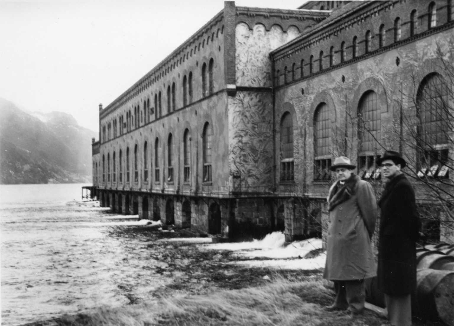 To menn foran stor murbygning ved vann.