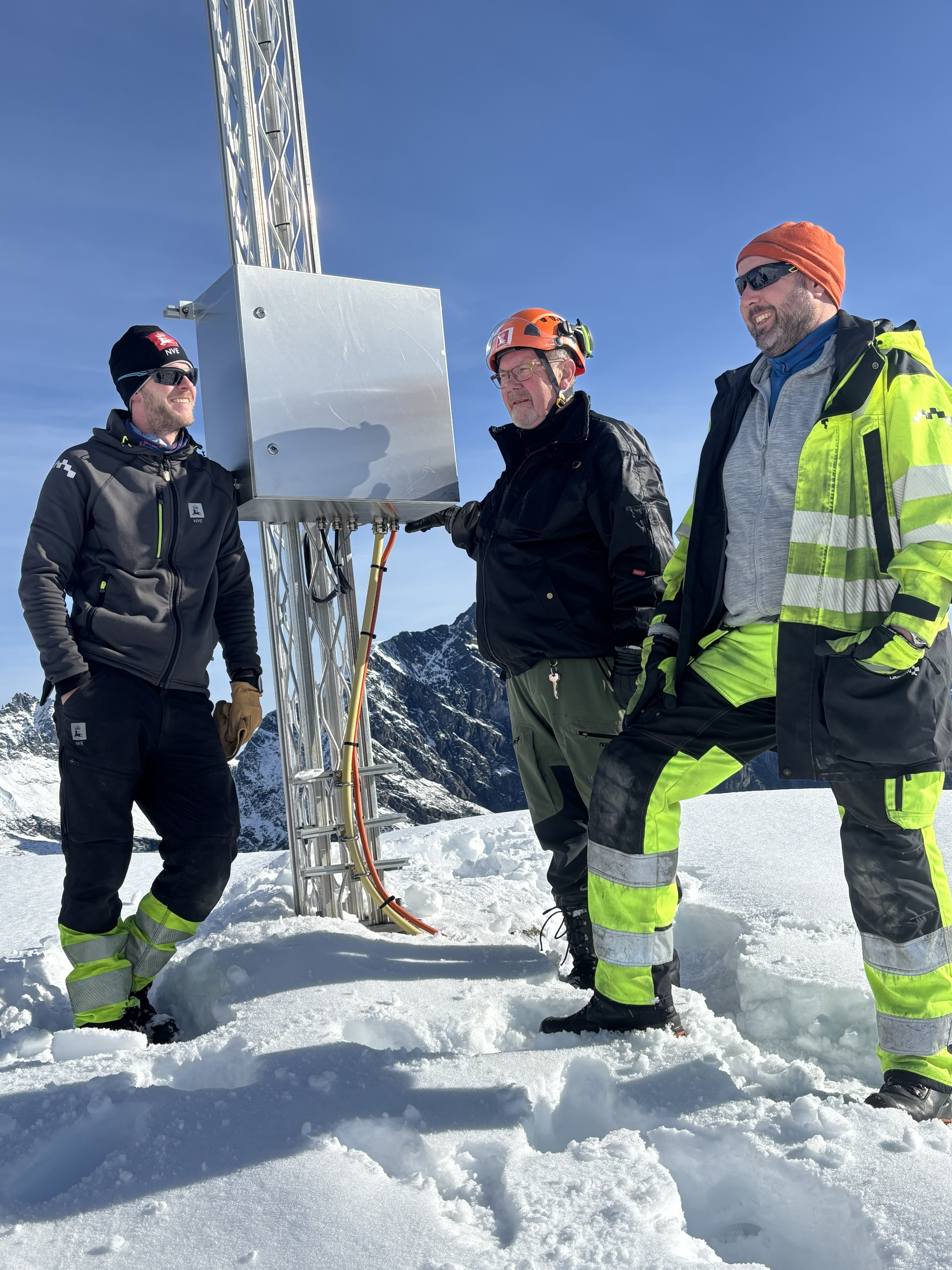 Tre menn står med arbeidsklær i snø ved en mast med en kasse på. Det er fjell i bakgrunnen.