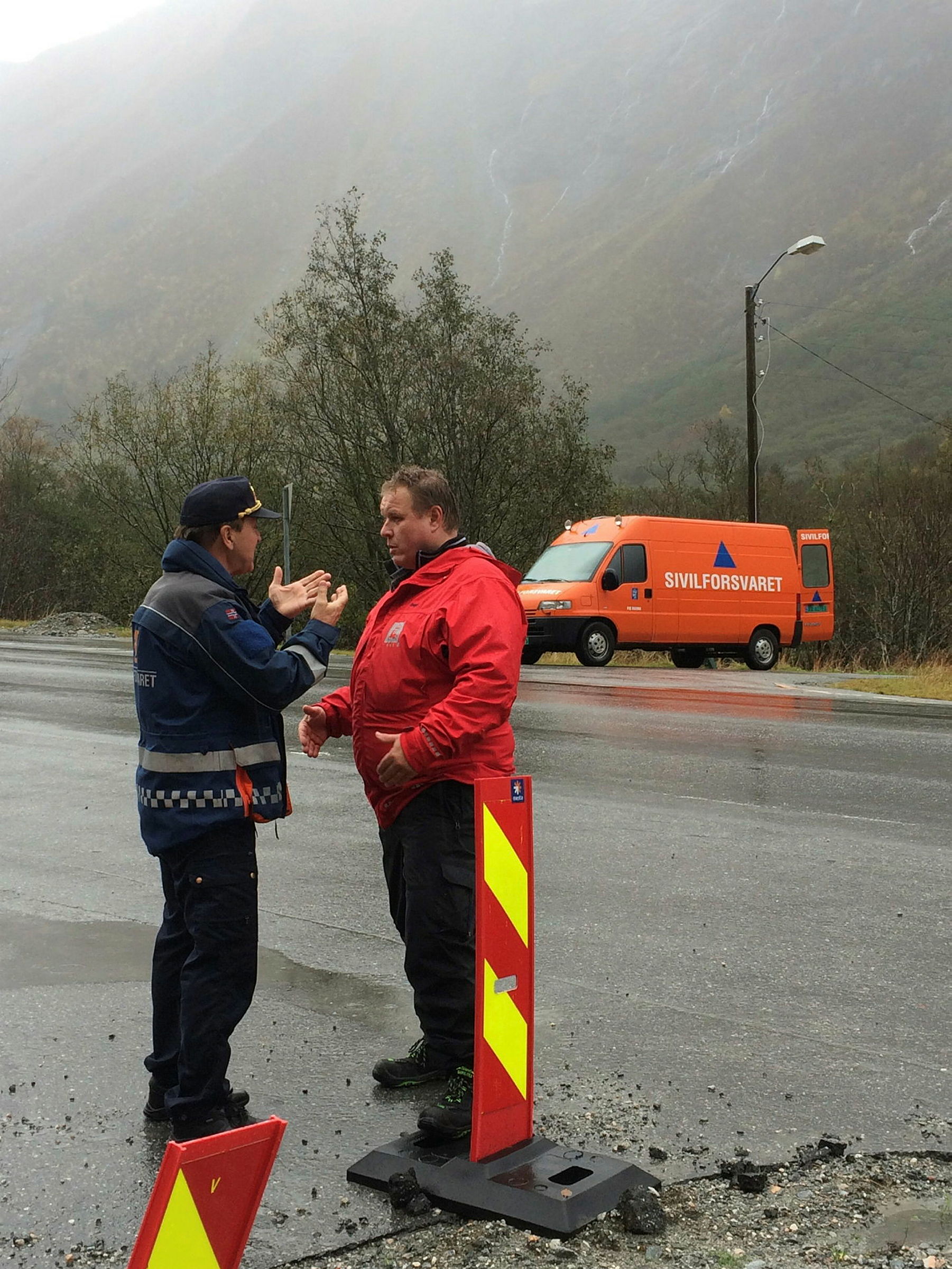 En politimann står i en diskusjon med en mann i rød jakke. I bakgrunnen er den en oransje varebil med logoen til sivilforsvaret. Det er høye fjell i rundt dem.