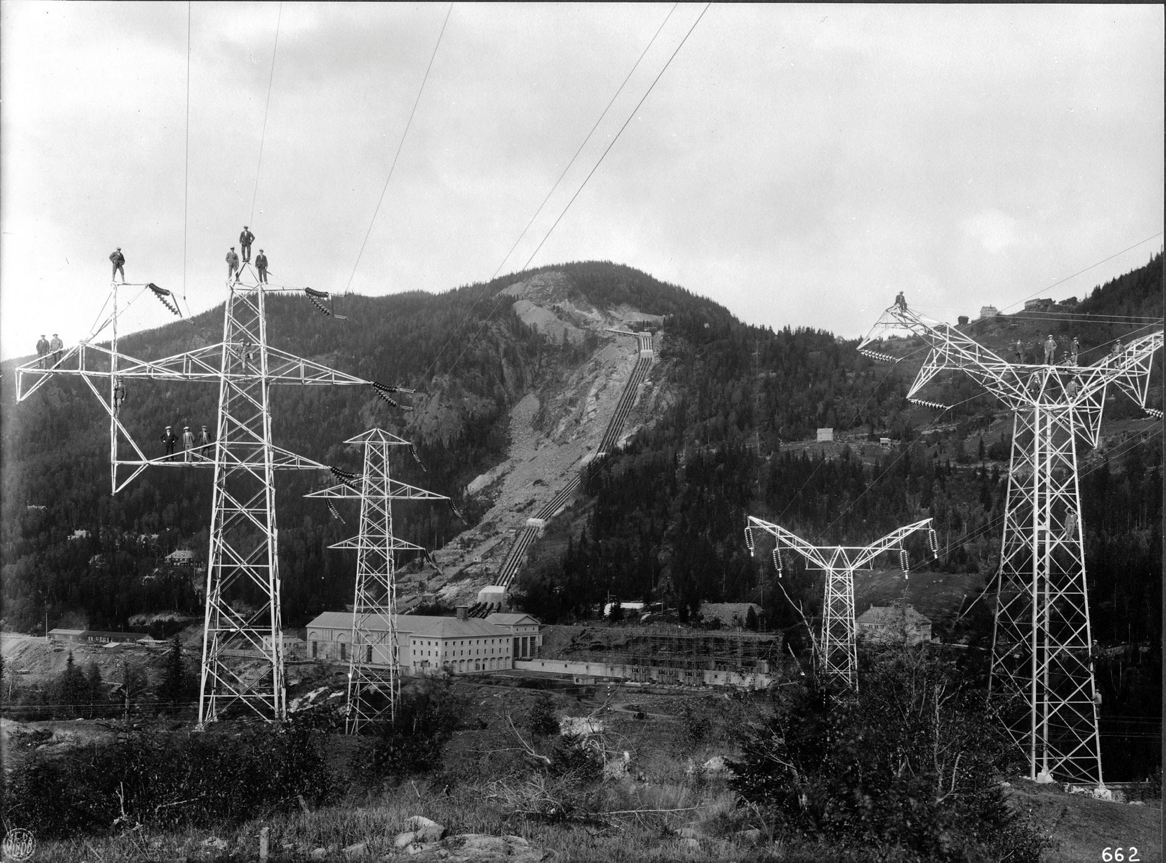 Mennesker står på toppen av to høye kraftmaster foran et fjell med en kraftstasjon i bakgrunnen