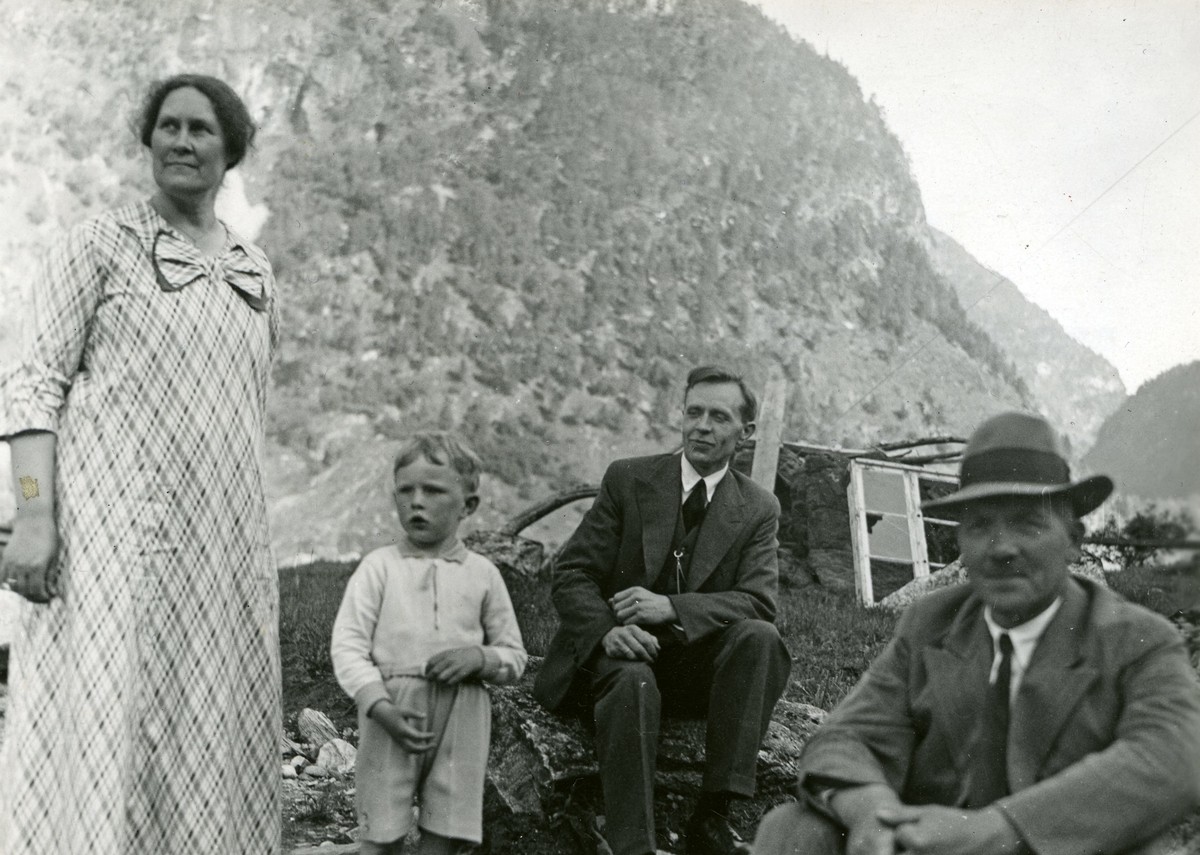 Fire mennesker ser mot samme punkt bak kamera. Fra venstre en dame som står, et lite barn, en mann sitter i dress og til høyre en mann i dress med hatt