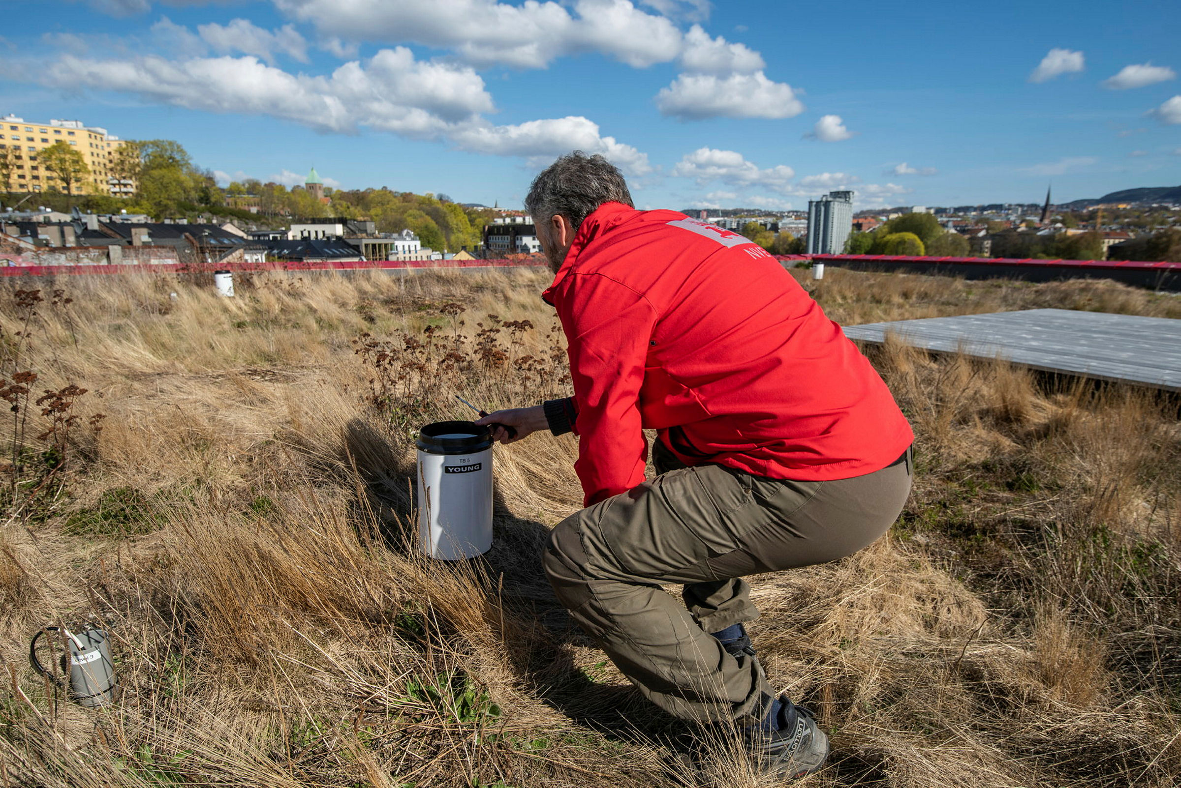 Mann i rød jakke undersøker et instrument på en gresskledt flate. Omgitt av bymiljø. 