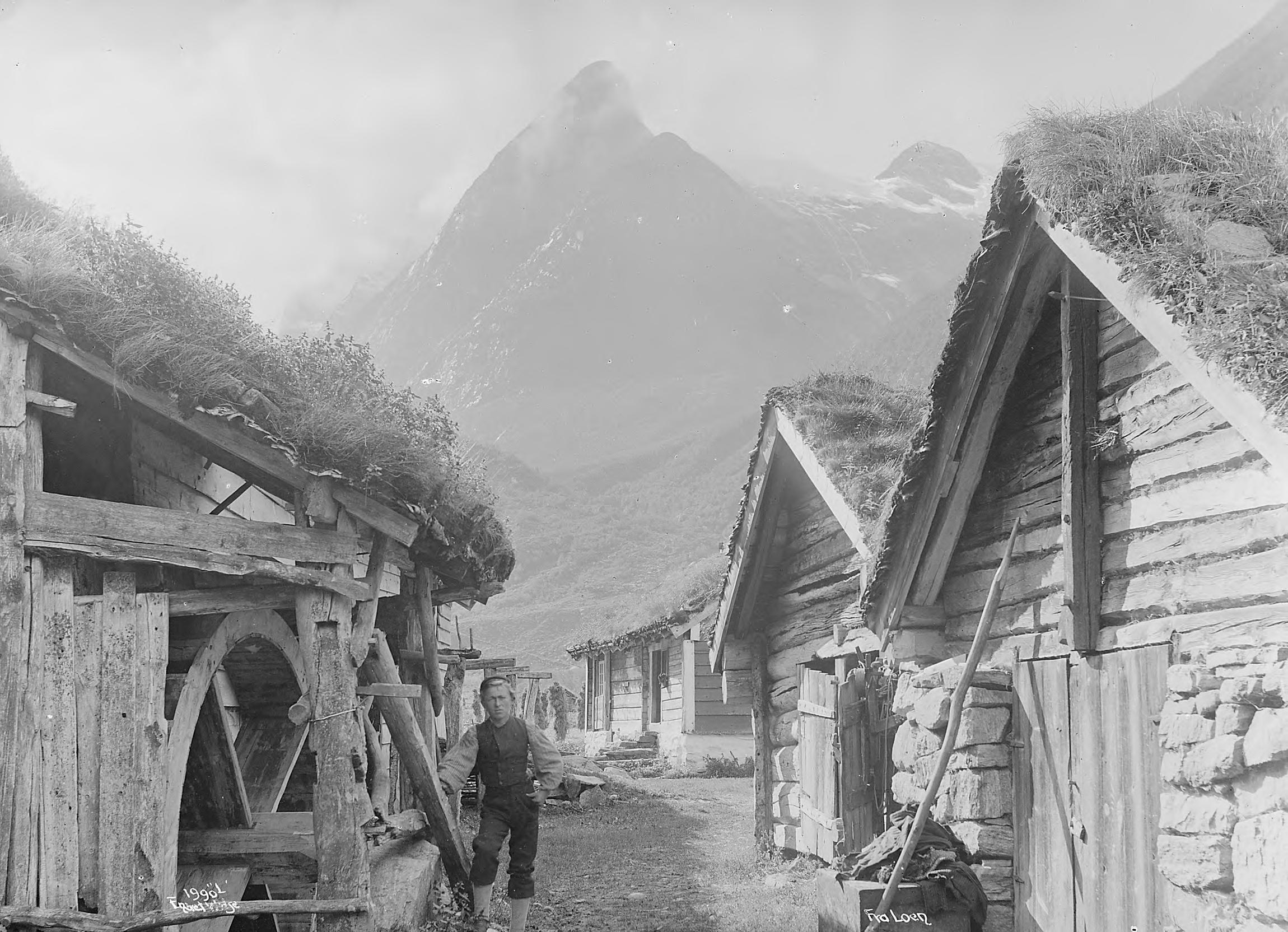 Gammelt bilde som viser en mann som står ved et vanndrevet treskehus og på andre siden ligger to fjøs og bakerst et hus til å bo i. I bakgrunnen ser vi et stort bratt fjell. 
