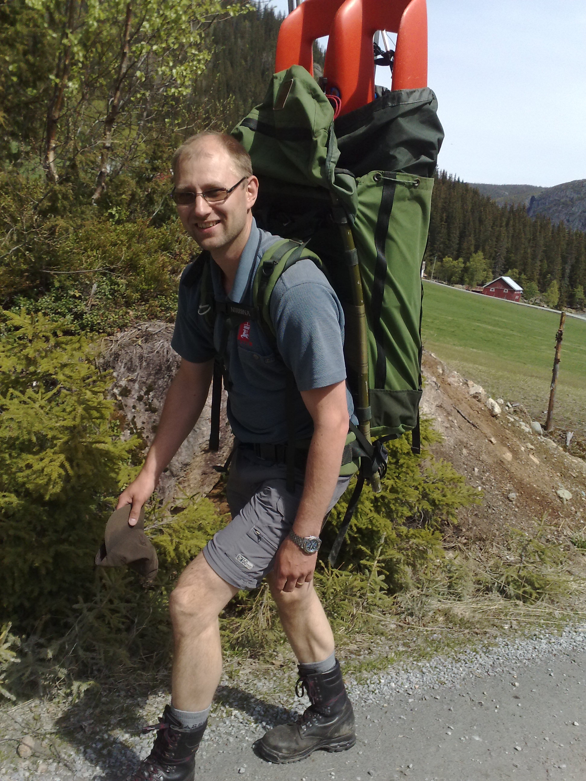 Mann med stor ryggsekk med utstyr i. Grønne omgivelser, med rødt hus i bakgrunnen.