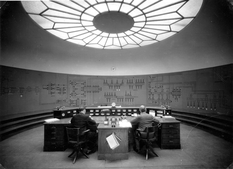 To menn sitter ved stort skrivebord med instrumenter i et kontrollrom. 