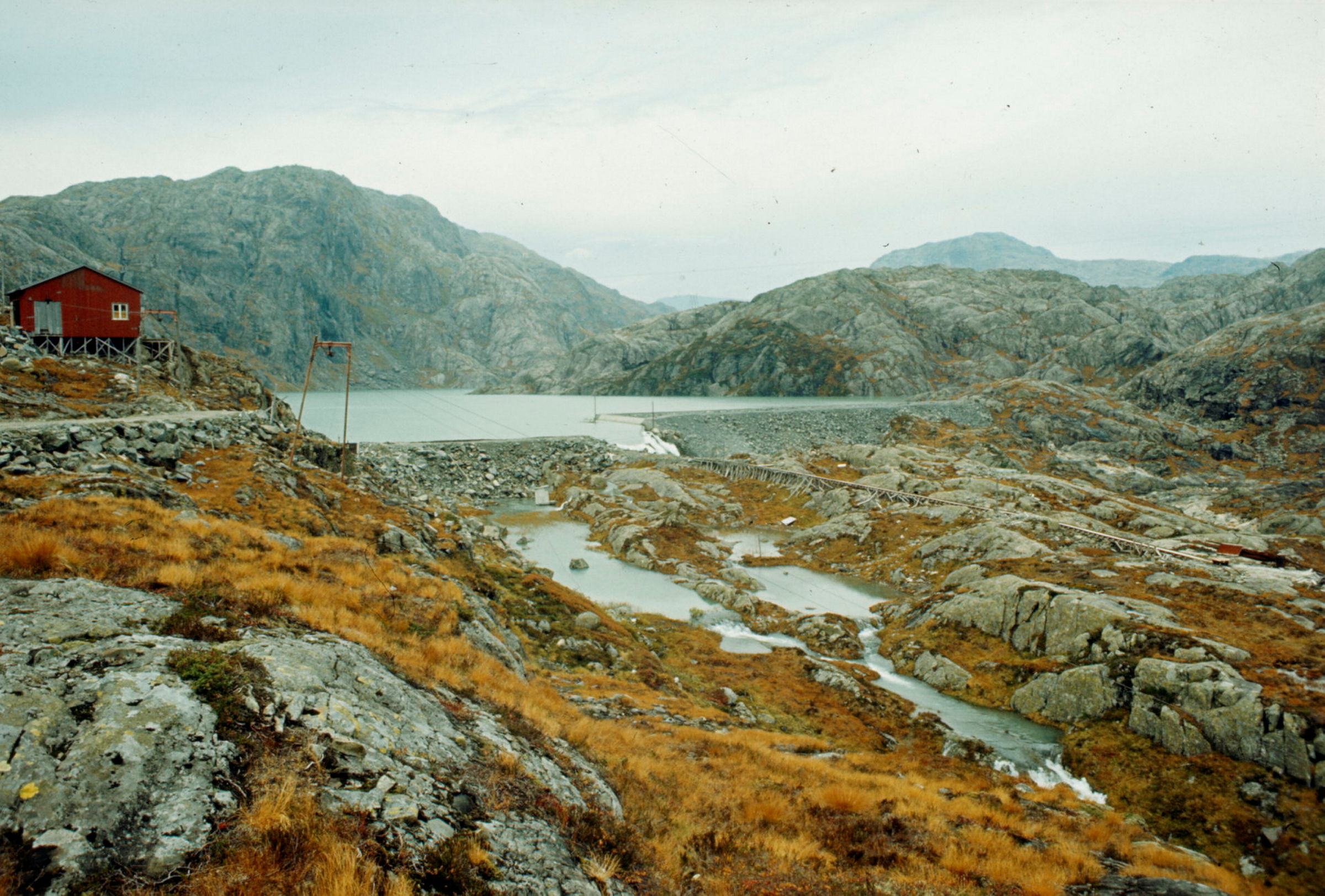 Landskapsbilde i høstfarger. Fjellparti med en dam som har demmet opp vann i et magasin. Kraftlinje midt i bildet. Rød trebygning til venstre.