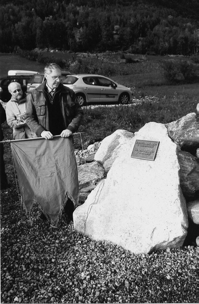 En mann som holder en vimpel står ved siden av en sten med en plate med skrift på. Bak han står flere menn og ser på stenen.