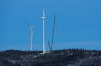 Bilde av en kran som heiser opp en turbinvinge til en vindturbin som er under bygging. Ved siden av til venstre står en vindturbin med alle tre turbinbladene festet til tårnet.