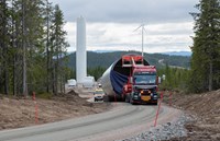 Lastebil, merket med bred last, som frakter en stor del til en vindturbin på en grusvei i et skoglandskap. 