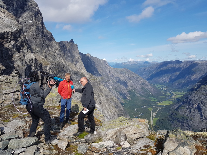Tre menn står ved en bratt fjellkant på et høyt fjell. En står med et stor kamera på skulderen og filmer de to foran kamera. En mann har en blå mikrofon i hånda og den andre har på en rød arbeidsjakke.