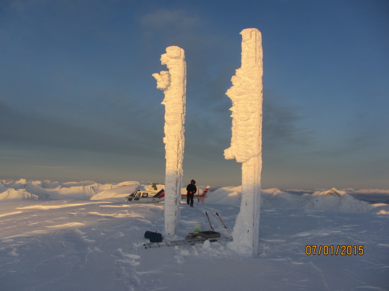 To snødekte stopler står i et snødekt fjellandskap. I bakgrunnen står et helikopter og en mann i vinterklær går mot stolpene.