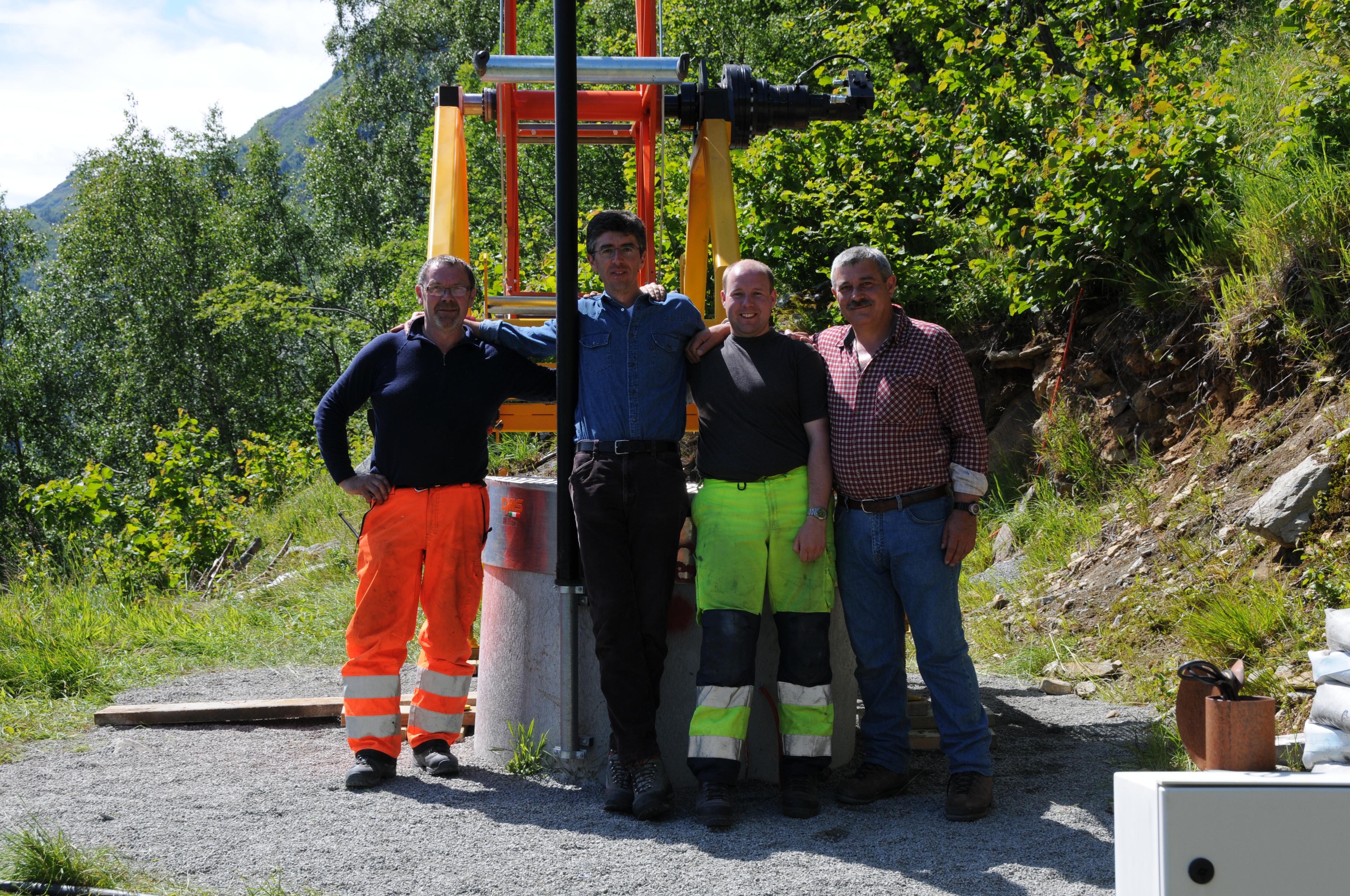 Fire menn står med armene rundt hverandre foran et høyt verktøy med grønt landskap i bakgrunnen.