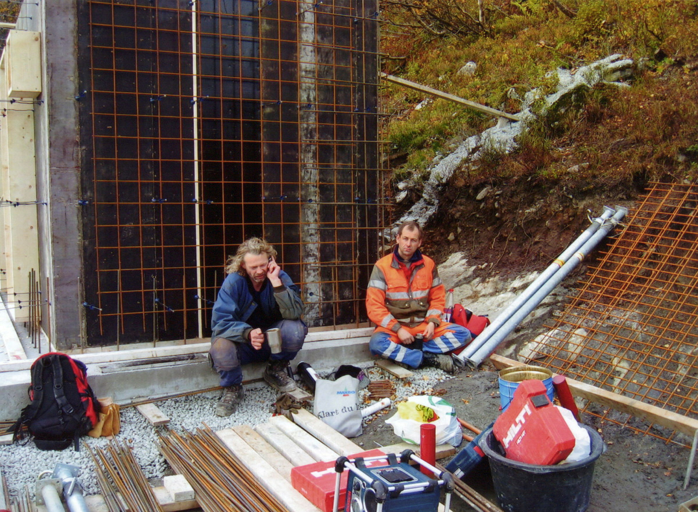 To menn i arbeidsklær sitter på en byggeplass og drikker fra en termokopp.