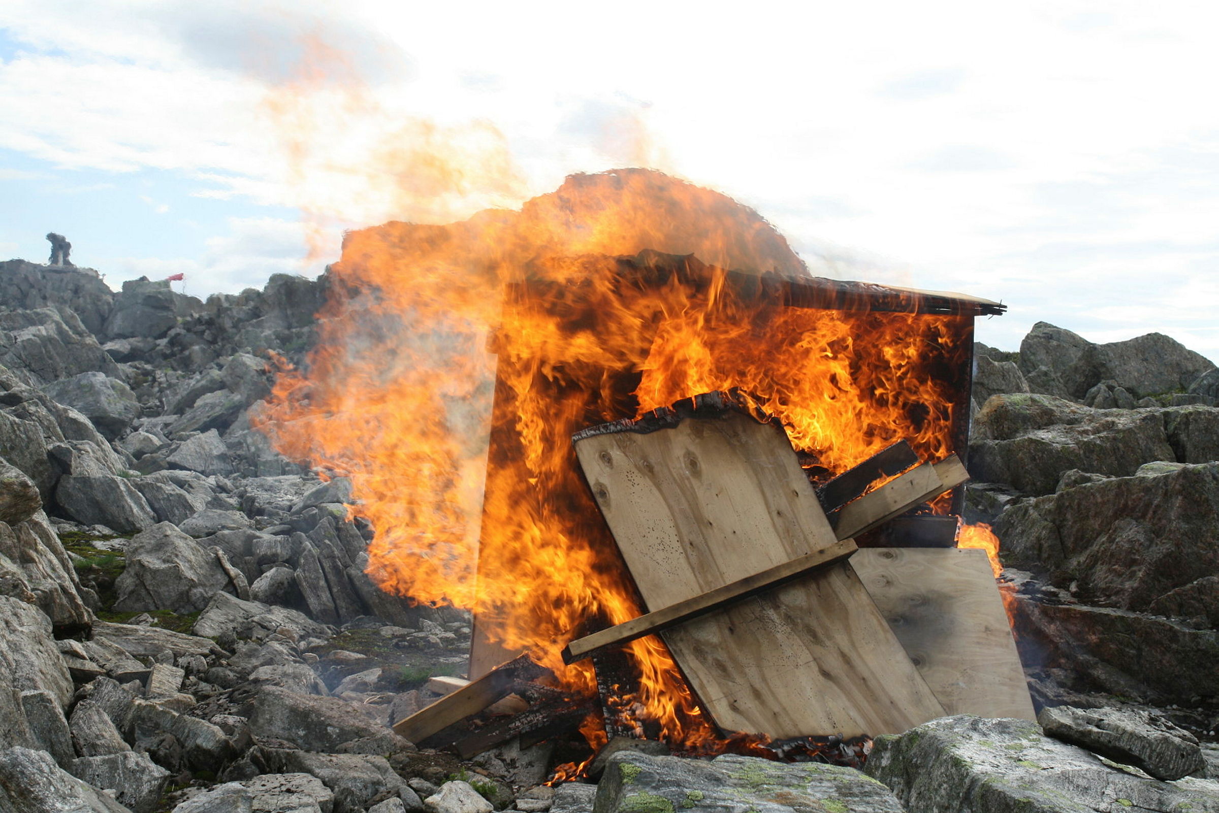 Et lite enkelt trehus står i flammer omringet av store steiner.
