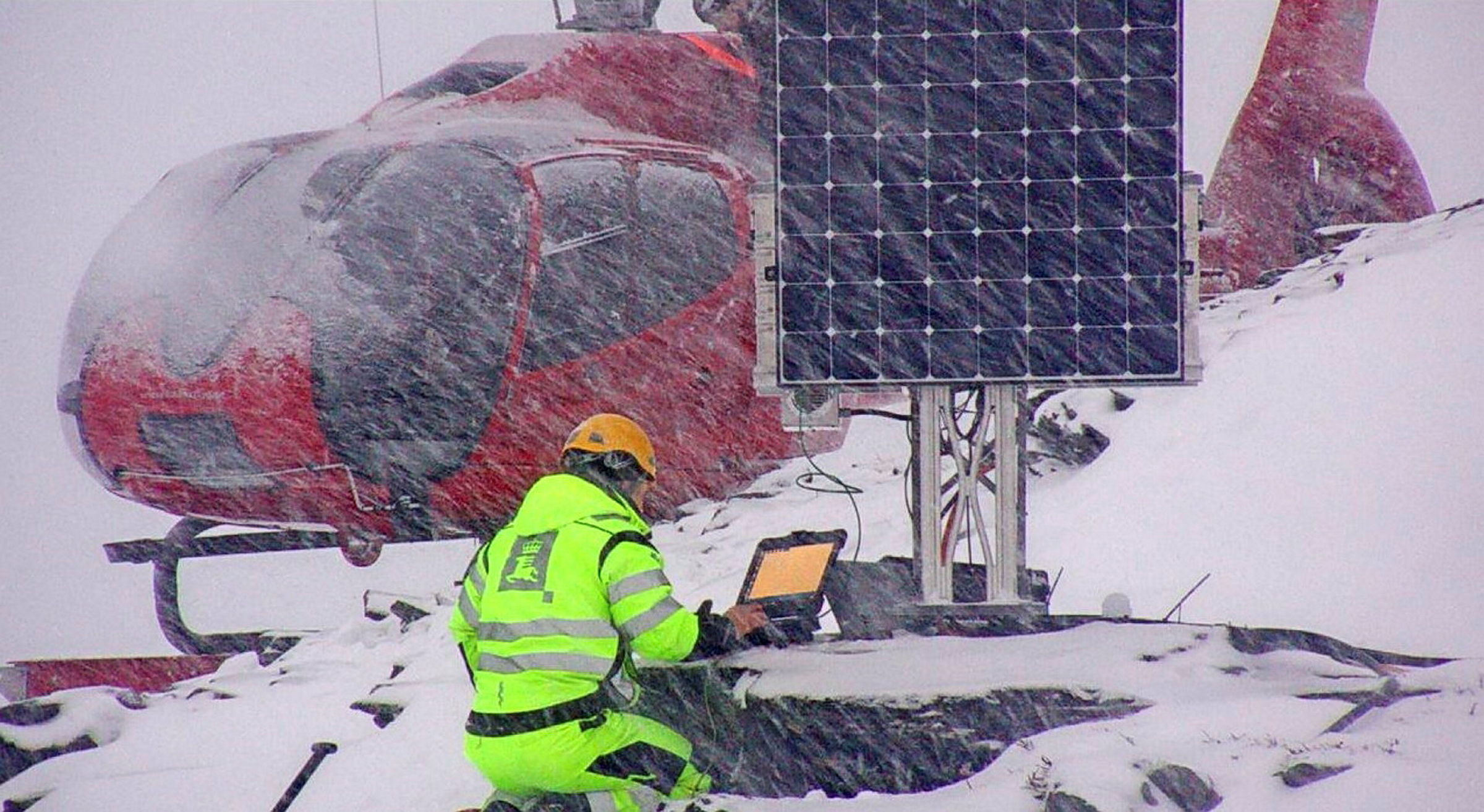 En mann i gule arbeidsklær og hjelm sitter foran en pc-skjerm ved et instrument. Bak ham står et rødt helikopter og rundt han er det kraftig snøvær.
