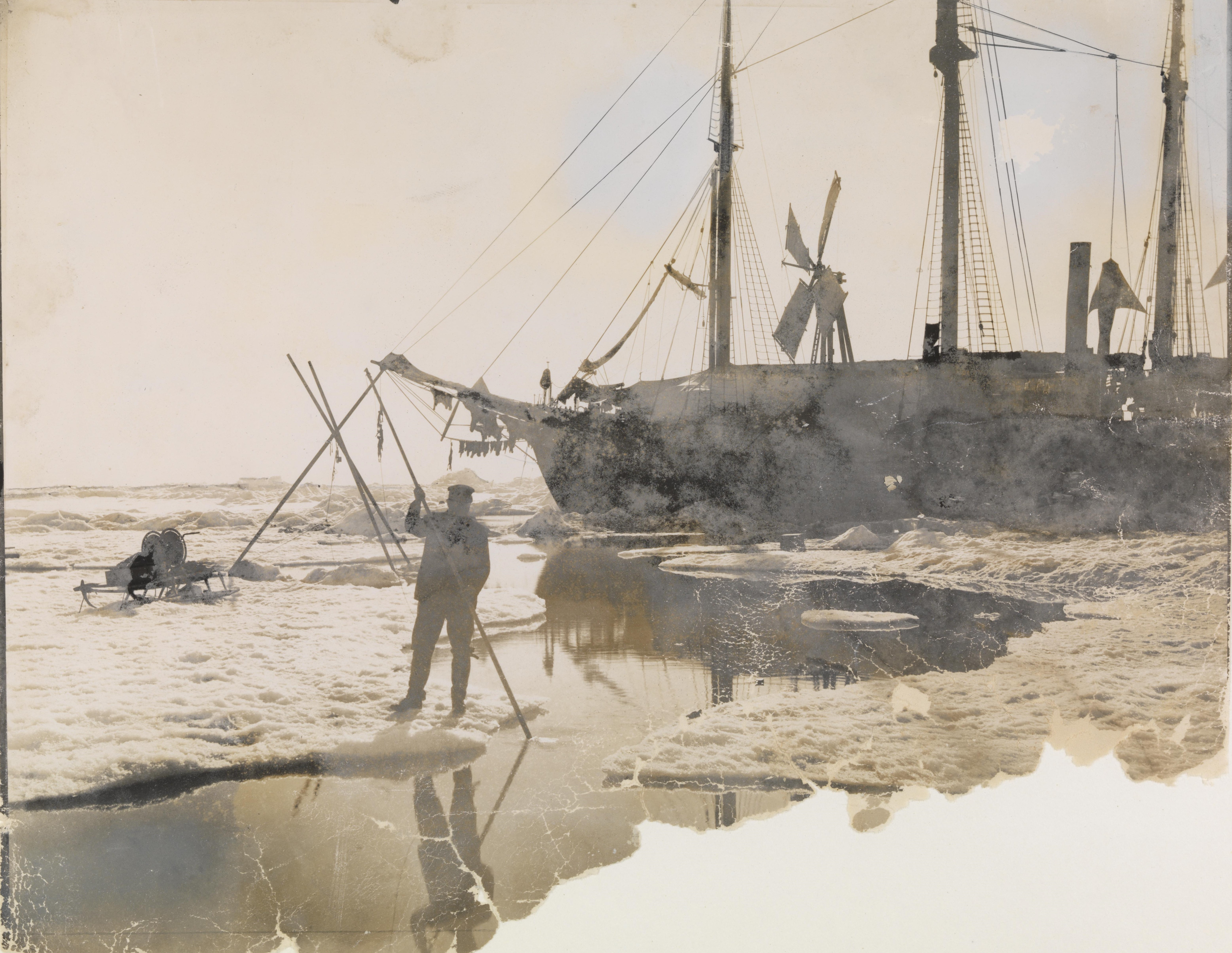 Foto av en mann ved en åpen isråk, foran polarskuten Fram som har en vindmølle montert på dekk. 