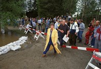 Kvinne i gul kåpe følge med flere personer. Til venstre, vann og sandsekker. Til høyre, mennesker som ser på følget. 