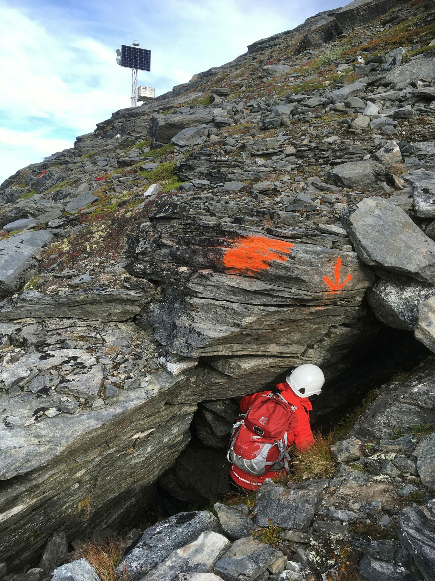 Person med sekk på ryggen smyger seg ned i fjellsprekk. Inngang markert med oransje spraymaling på berget.