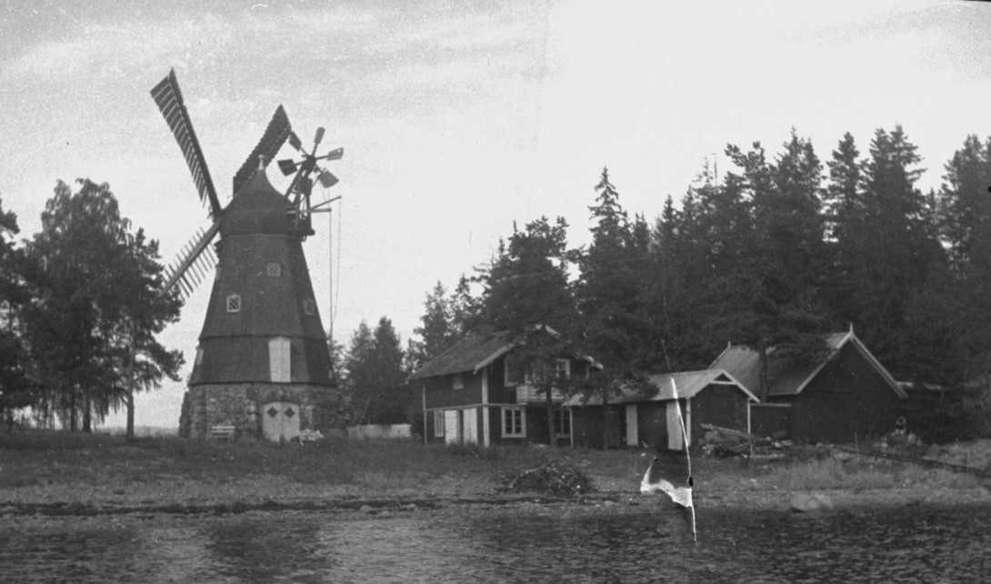 Eldre bilde som viser en hettevindmølle ved siden av et lite tun med hus nær stranden.