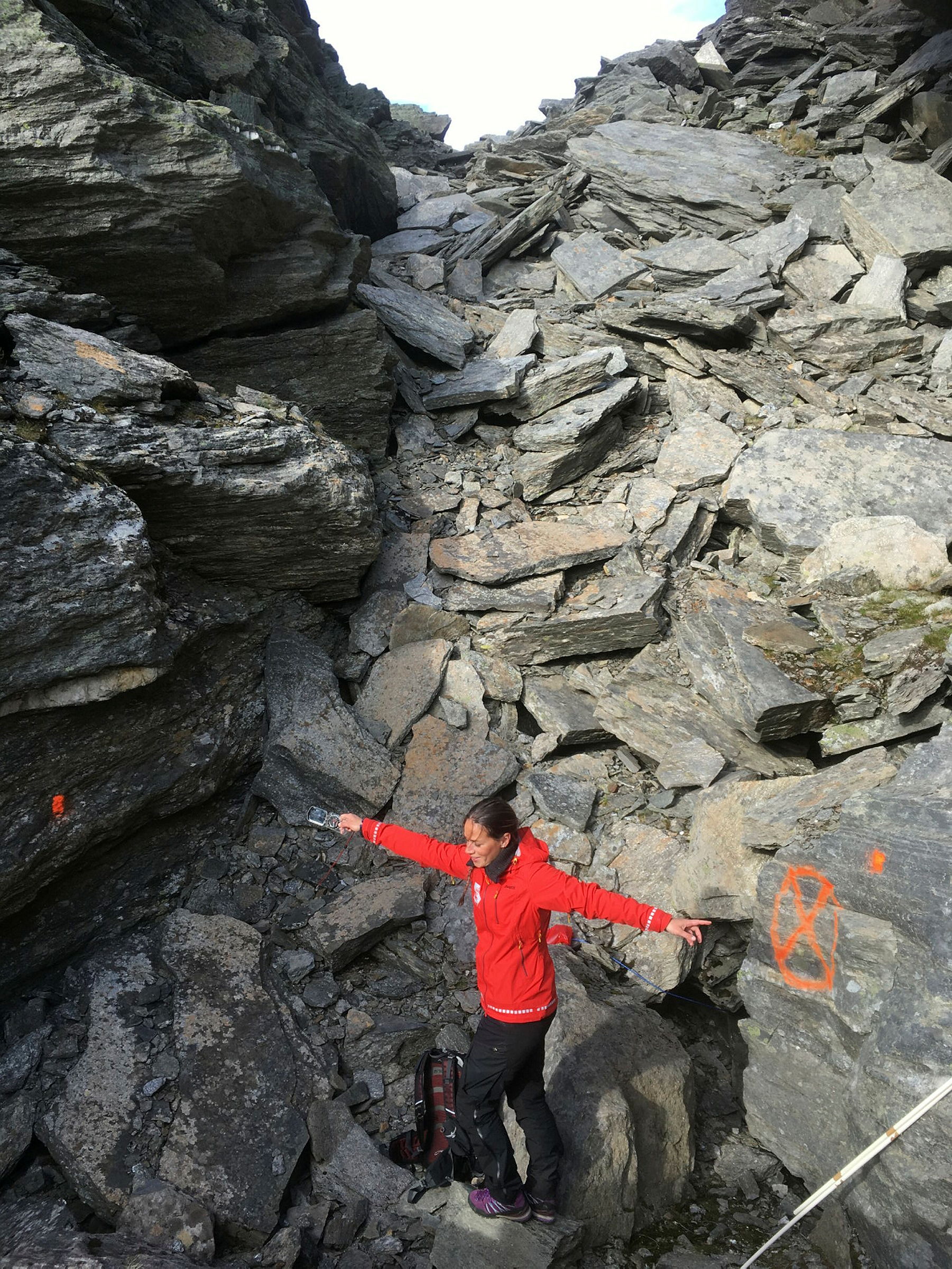 Person i rød NVE jakke viser med utstrakte armer målepunkter for fjellsprekken markert med oransje spraymaling på berget.
