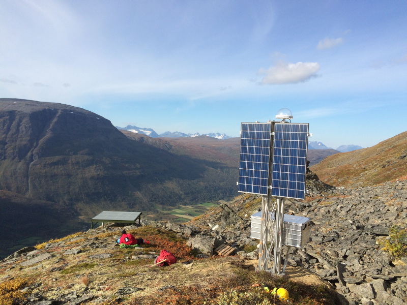 Et solcellepanel står på toppen av et fjell med grønt fjordlandskap i bakgrunnen.