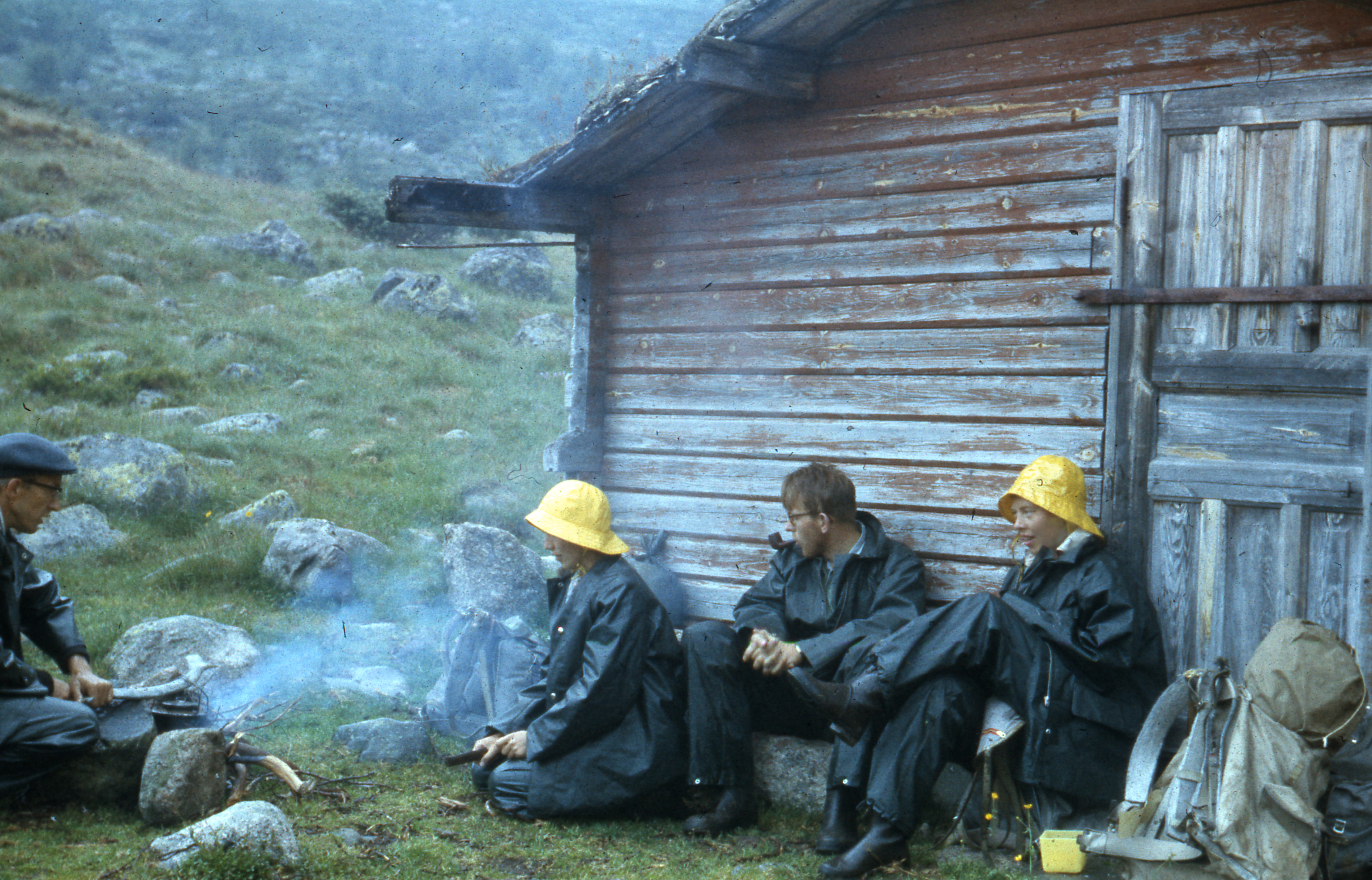 Fire personer sitter i regntøy rundt et bål, og med ryggen mot en tømmervegg.  
