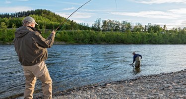 To menn der den til venstre står på land og dra inn fisk med fiskestang. Den andre i vadebukser står i elven.
