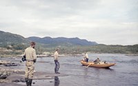 To personer står i vannkanten og ser mot to personer i båt som forsøker å legge til. 