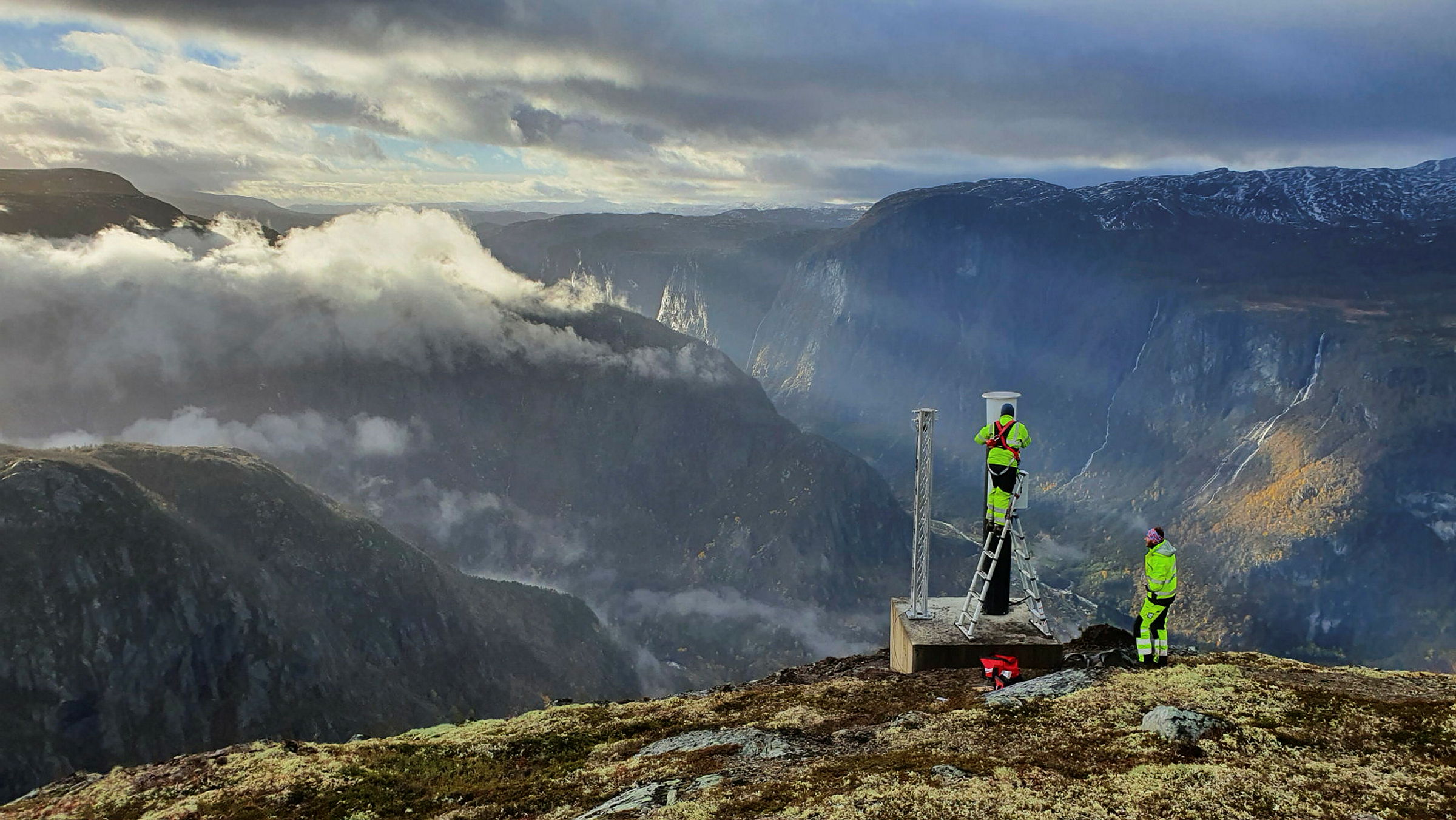 To menn i gult arbeidstøy står ved et apparat oppe på en fjelltopp. I bakgrunnen er det store fjell.